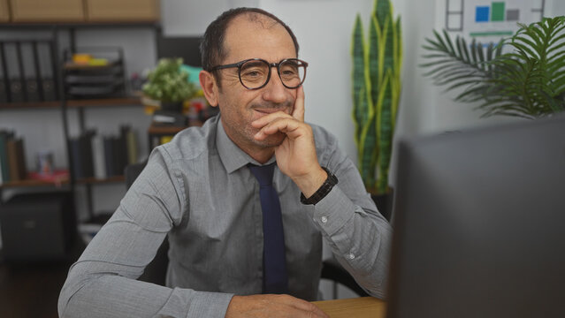 Hispanic middle-aged man in glasses working in a modern office, focused intently on his computer screen with a thoughtful expression.