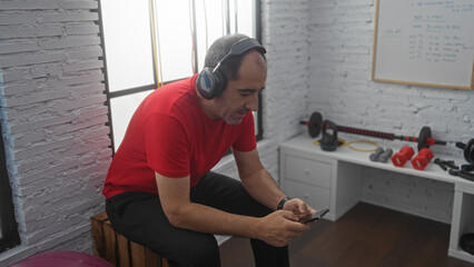 A middle-aged hispanic man in a red shirt and black pants is sitting in a gym, wearing headphones and looking at his phone.