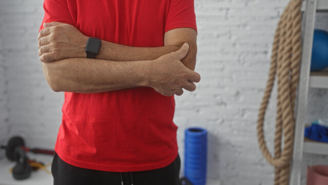 Hispanic man in red shirt touching elbow in indoor gym with workout equipment and ropes on the wall