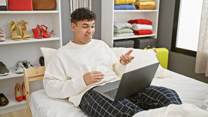 A young man using a laptop in his modern bedroom, casually dressed and interacting with someone online