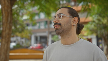 Young hispanic man with a moustache and glasses standing outdoors in a city park, looking pensive with urban buildings in the background