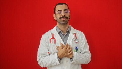 Handsome young hispanic man wearing a lab coat and stethoscope stands with hands on chest against a vibrant red background with a content expression