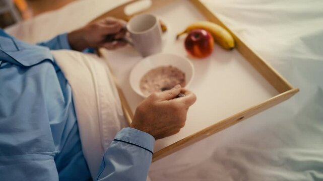 Aged male eating breakfast and drinking tea while lying in a nursing home bed