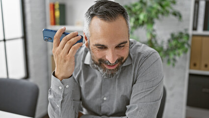 A smiling mature hispanic man with grey beard listens to a smartphone in an office setting.