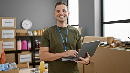 Smiling hispanic man with a beard working on a laptop in a warehouse full of cardboard boxes.