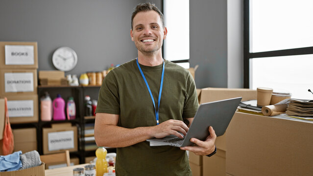 Smiling hispanic man with a beard working on a laptop in a warehouse full of cardboard boxes.