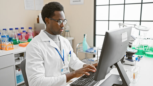 A focused african american man wearing a lab coat works on a computer in a bright laboratory setting.