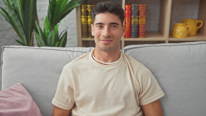 Handsome young hispanic man smiling while relaxed on a grey sofa in a cozy living room interior.