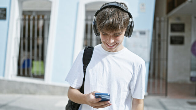 A smiling young caucasian teen boy listens to music on headphones while using a smartphone on a sunny city street.