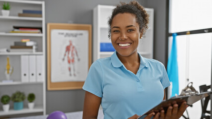 Smiling woman holding clipboard in a physical therapy clinic interior with anatomical posters and exercise equipment.