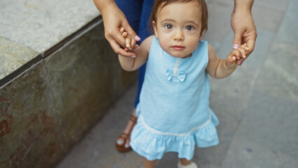 Fototapeta premium Woman holding toddler's hands on urban street, showcasing love, togetherness, and family in an outdoor city setting with a young child in a blue dress