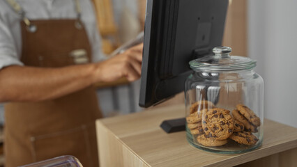 Young man working in a bakery shop with cookies in a glass jar and using a computer in an indoor setting