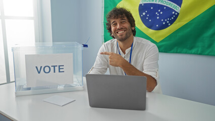 Man sitting indoors pointing at a voting box in front of a brazilian flag in a college setting, suggesting participation in an election.