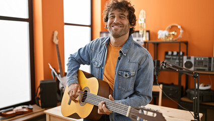 Handsome hispanic man playing guitar smiling in a music studio