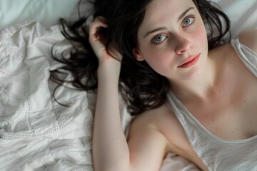 Naklejka premium A woman with long dark hair lies in bed, her eyes gazing upwards as the morning light illuminates her face