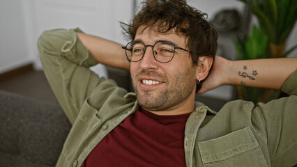 Obraz premium Relaxed young man with glasses and a beard smiling in a cozy indoor setting, portraying casual home comfort.