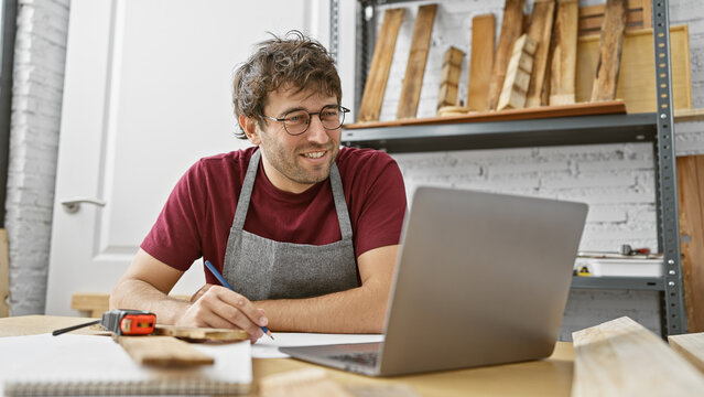 A smiling man wearing glasses and a workshop apron works on a laptop in a sunny carpentry studio surrounded by wooden planks.