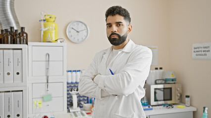 Confident hispanic man with beard in white lab coat standing arms crossed in medical clinic laboratory