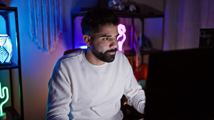 Hispanic man with beard focused on computer screen in a neon-lit gaming room at night.