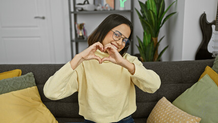 Obraz premium A cheerful young hispanic woman makes a heart shape with her hands whilst sitting on a sofa in a cozy living room.