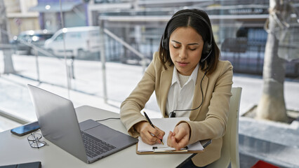 A focused hispanic woman in a beige blazer taking notes with a laptop on a white table against a blurred background.