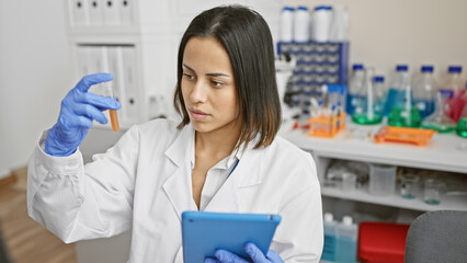 A focused young woman scientist examining a test tube in a modern laboratory while holding a tablet.