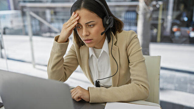 Stressed young hispanic woman in a headset sits at her desk with a laptop, embodying the challenges of customer service.
