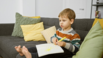 Blond toddler boy drawing indoors on a couch with colorful pillows in a cozy living room setting