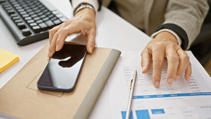 Middle-aged woman analyzing financial documents in office, with smartphone and keyboard on desk.