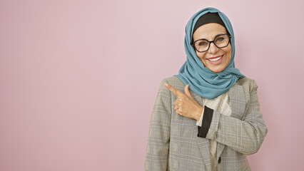Smiling middle-aged hispanic woman in a hijab, pointing to her side against a pink wall.