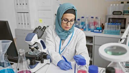 A mature woman scientist in hijab meticulously taking notes in a laboratory setting.