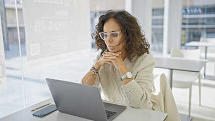A thoughtful hispanic businesswoman works on a laptop in a modern office setting, showcasing technology, professionalism, and focus.