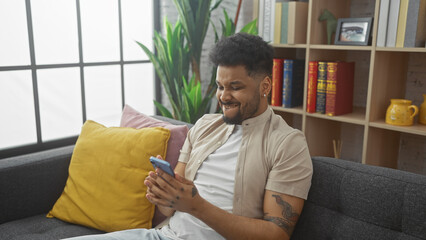 African american man smiling at smartphone in cozy living room with books and plants.