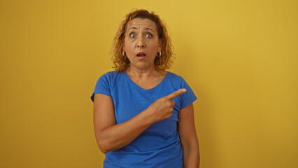 A middle-aged hispanic woman with curly hair in a blue shirt stands isolated against a yellow background, appearing surprised while pointing to the side.