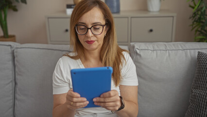 Middle-aged woman wearing glasses and using a blue tablet while seated on a grey sofa in a modern living room of an apartment.