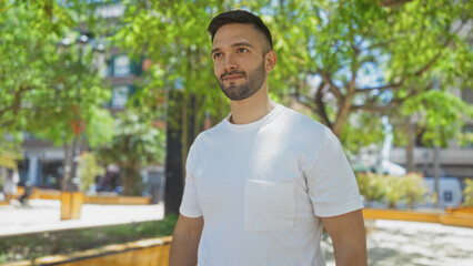 Handsome young hispanic man with a beard wearing a white shirt, standing outdoors in a city park with greenery and buildings in the background.