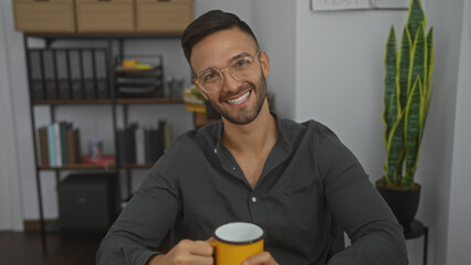Handsome young hispanic man with a beard and glasses sitting in an office holding a yellow mug while smiling, with shelves and indoor plants in the background.