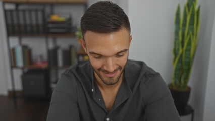 Fototapeta premium Handsome young hispanic man with a beard smiling indoors at his workplace, surrounded by office supplies and a plant in the background.