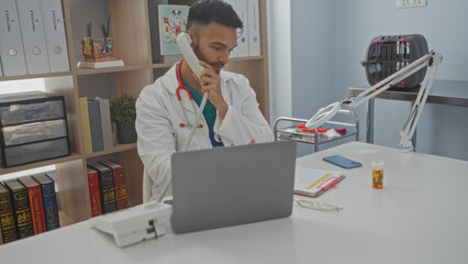Obraz premium Young hispanic man in a veterinary clinic talking on the phone while working on a laptop in an indoor office setting with medical equipment and shelves in the background.