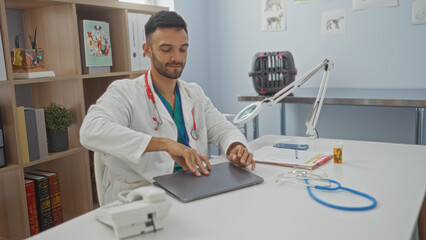 Young, hispanic man with a beard working in a veterinary clinic, sitting at a desk with a stethoscope, notebook, and medication bottle, surrounded by books and animal posters.