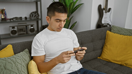Handsome young hispanic man using smartphone on couch at home with guitar background