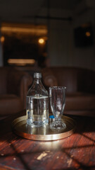 An empty transparent bottle and glass on a golden tray placed on a wooden table in a cozy dimly lit room.