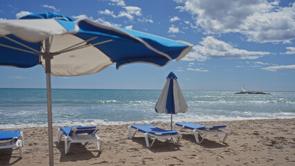 A tranquil seaside scene with sun loungers and umbrellas on a sandy beach against a backdrop of sparkling ocean water and a clear sky.
