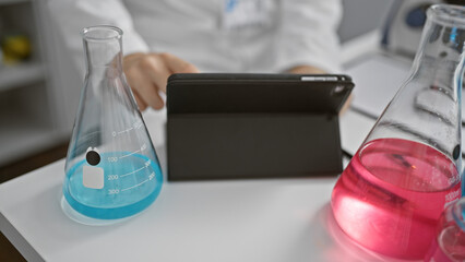 A young man in a laboratory analyzes results on a tablet, surrounded by beakers with blue and red chemicals