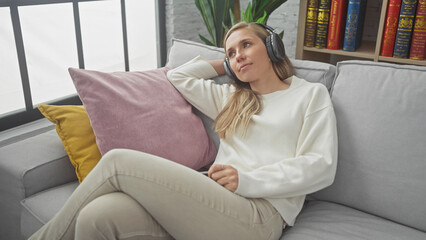 A pensive young woman enjoys music on headphones while relaxing alone on a sofa in a cozy living room.