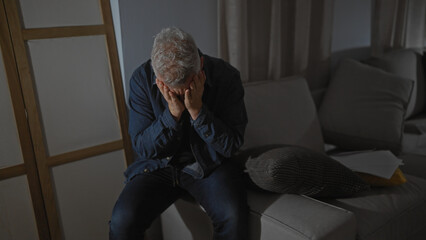 Distressed man sitting indoors covering face with hands, showing sorrow in a dimly lit room