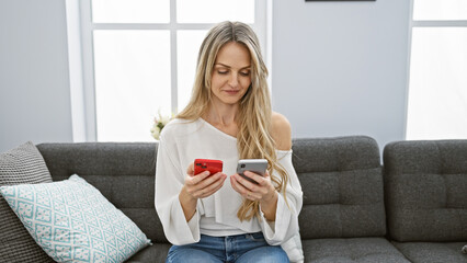 A young blonde woman compares smartphones while sitting comfortably in her modern living room.