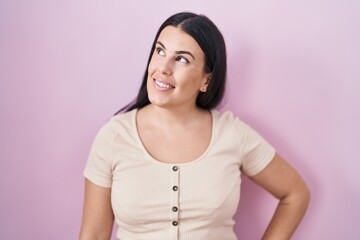 Fototapeta premium Young hispanic woman standing over pink background looking away to side with smile on face, natural expression. laughing confident.