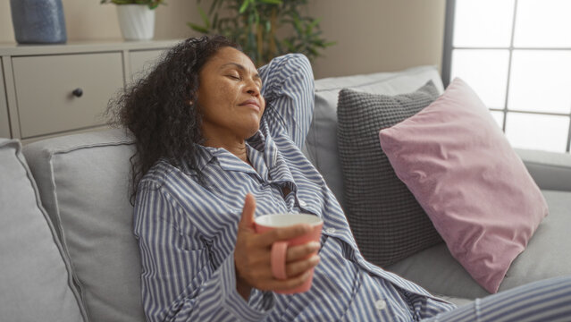 Woman relaxing with a coffee cup on a sofa in a living room, wearing striped pajamas, with eyes closed and sunlight coming through a window, creating a peaceful and serene atmosphere