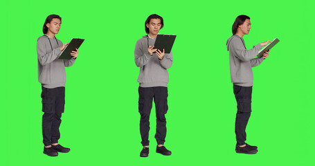 Asian adult reading information on files, thinking about annotations on clipboard papers or taking notes for finalization. Young guy writing on documents, standing against greenscreen.